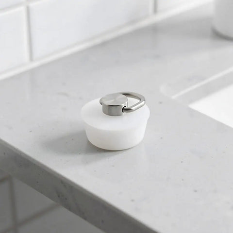 White Drain Stopper with Stainless Steel Ring, Silicone Seal Plug resting on a marble countertop by the sink.