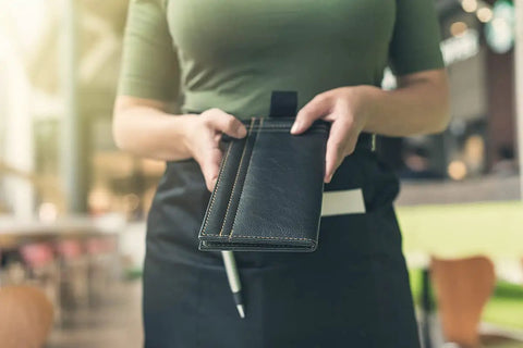Server wearing a black apron holding a checkbook in a restaurant, part of 12 Pack Server Aprons With 3 Pockets For Waitstaff, Water Resistant Half Aprons For Women And Men.