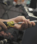 Hands holding All In One Multi Bit Screwdriver And Tap Tool with yellow-black handle in a workshop setting.