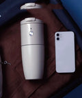 Compact Portable Coffee Maker With Built In Automatic Bean Grinder For Home Desk Or Road Trip next to a smartphone in a suitcase.