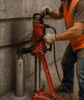 Diamond core drill on red stand with silver 3-inch bit against concrete wall; worker in orange safety vest.