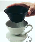 Hand holding a black ceramic dripper above a white ceramic cup, showcasing the Prime Pick Japanese Ceramic Coffee Dripper With Reusable Filter And Ultra Fine Pores And Eco Friendly Hand Pour Setup For Straight Up Smooth Daily Brews Of Coffee Tea Or Water.