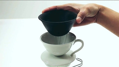 Hand holding a black ceramic dripper above a white ceramic cup, showcasing the Prime Pick Japanese Ceramic Coffee Dripper With Reusable Filter And Ultra Fine Pores And Eco Friendly Hand Pour Setup For Straight Up Smooth Daily Brews Of Coffee Tea Or Water.