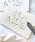 Close-up of a grey foil quill pen writing gold script on a white wedding card.