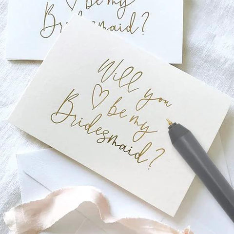 Close-up of a grey foil quill pen writing gold script on a white wedding card.