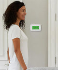 Woman near wall-mounted RTH7600D 7-Day Programmable Touchscreen Thermostat with Home and Away Temperature Settings, white finish.