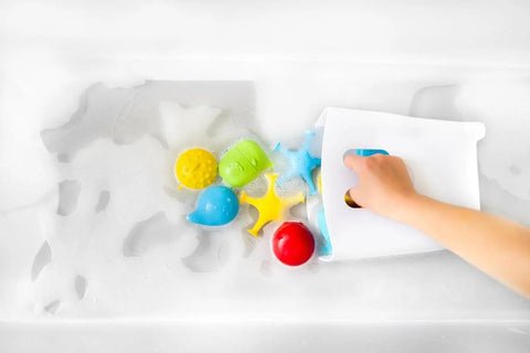 Child’s hand using the Freestanding Bath Toy Drying Bin With Quick Drain Base in soapy water to organize colorful bath toys.