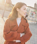 Profile view of a woman wearing an orange coat, standing outdoors in sunlight, background of city scenery.