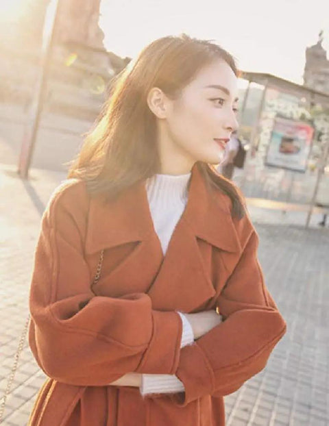 Profile view of a woman wearing an orange coat, standing outdoors in sunlight, background of city scenery.