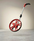 Hella Portable Foldable Distance Measuring Wheel With Bright Lcd Screen And Built In Carry Strap Rolls Out Smooth For Contractors And Handyman Teams, featuring red wheel and silver handle.