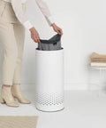 Person placing bag into 9 Gal White Laundry Hamper With Quick Drop Lid For Easy Toss In Ventilation Holes Corrosion Resistant Materials Hygienic And Discreet Laundry Basket For Bathroom Or Laundry Room