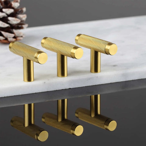 Three gold knurled cabinet T-pulls on a marble slab, reflecting under soft lighting.