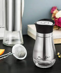 Go-To Salt And Pepper Shaker Set With Stainless Steel Caps And Glass Bottles on a table with spices and decor