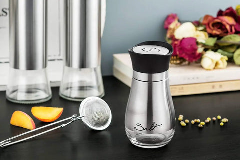 Go-To Salt And Pepper Shaker Set With Stainless Steel Caps And Glass Bottles on a table with spices and decor