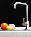 Stainless steel kitchen faucet with three fruits: orange, lemon, and apple resting on a countertop.