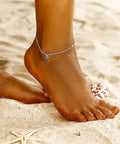 Person wearing silver anklet with turtle charm on sandy beach.