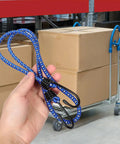 Person holding blue rubber bungee cords with buckle hooks in warehouse, boxes and dolly in background.