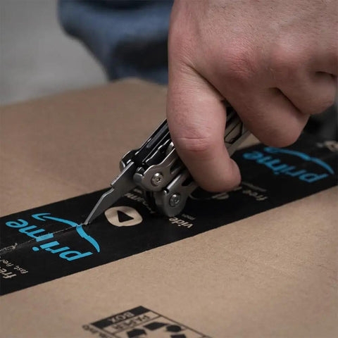 Close-up of hand using the 13 In 1 Stainless Steel EDC Multitool with knife blade to cut through packaging tape on a cardboard box.
