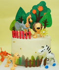 Jungle-themed cake with animal figurines, including an elephant, lion, giraffe, zebra, surrounded by red and brown picket fences.