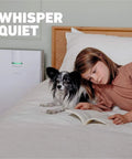 Front view of white Hathaspace HSP002 air purifier with green light beside bed; woman and dog reading a book.
