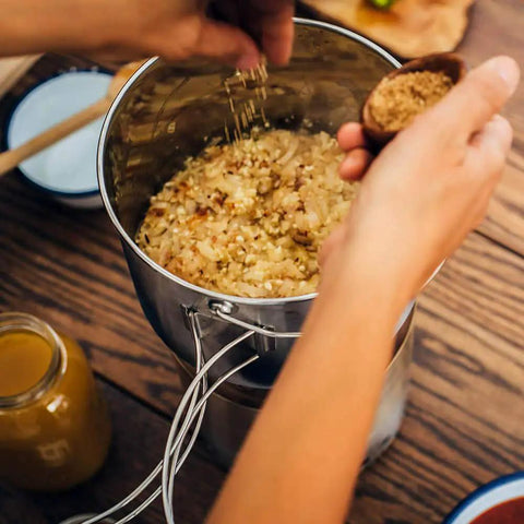 Hands adding spices to Pot 4000 Stainless Steel Camping Pot for outdoor cooking with foldable handles and rice inside.