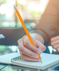 Person using yellow HB carpenter pencil with white eraser head for drafting and note-taking in a notebook on a glass table.