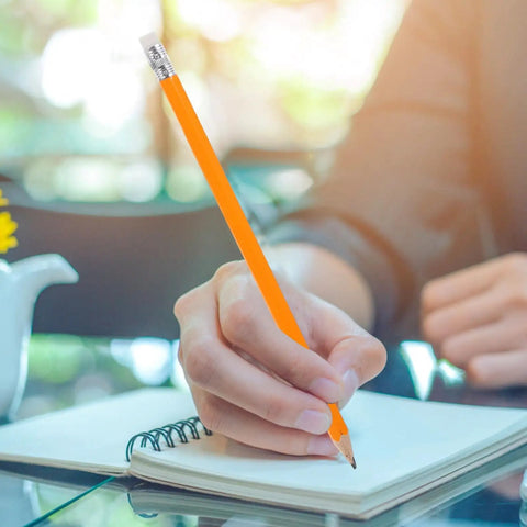 Person using yellow HB carpenter pencil with white eraser head for drafting and note-taking in a notebook on a glass table.