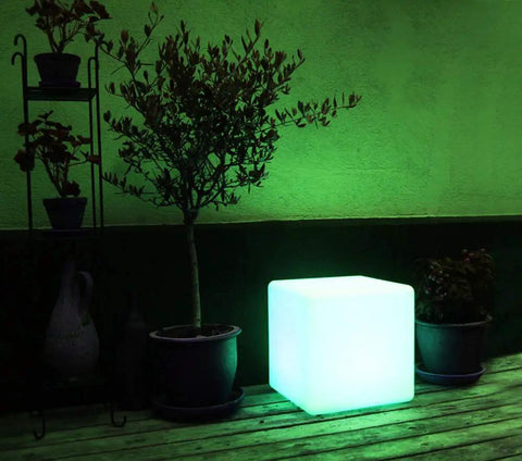 Rechargeable LED Color Changing Cube Light 8 Inch Dimmable with remote stands illuminated on a patio next to potted plants.