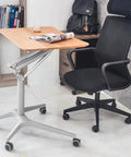 Hella Stable Mobile Standing Desk Featuring Instant Gas Spring Lift And Smooth Glide Rollers For A Total Game Changer Home Office Upgrade Or Flexible Workspace Setup In Clean Black next to a black office chair.