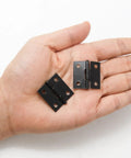 Two solid black one inch metal hinges in a hand, part of 24 Pack Of Solid Black One Inch Metal Hinges With Included Fasteners Quick Hook Up For Drawers Cabinets And Small Craft Boxes Built Tough For Heavy Duty Jobs.