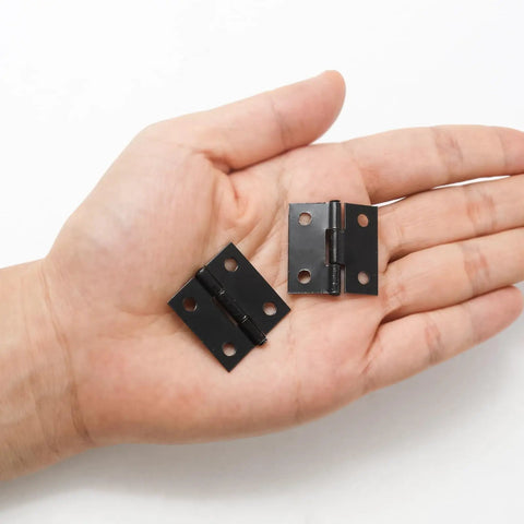 Two solid black one inch metal hinges in a hand, part of 24 Pack Of Solid Black One Inch Metal Hinges With Included Fasteners Quick Hook Up For Drawers Cabinets And Small Craft Boxes Built Tough For Heavy Duty Jobs.