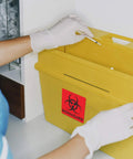 Hand in gloves placing syringe into yellow bin with Heavy Duty 12 Pack Biohazard Stickers 6X6 Inches on it.