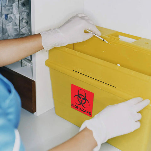 Hand in gloves placing syringe into yellow bin with Heavy Duty 12 Pack Biohazard Stickers 6X6 Inches on it.