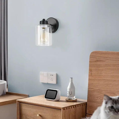 One Light Matte Black Bathroom Vanity Sconce with Clear Glass Shade over nightstand in room view.