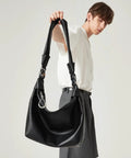 Person holding a large black leather handbag with a wide strap and silver hardware, wearing a white shirt and grey pants.