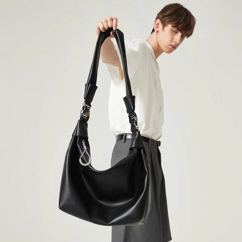 Person holding a large black leather handbag with a wide strap and silver hardware, wearing a white shirt and grey pants.