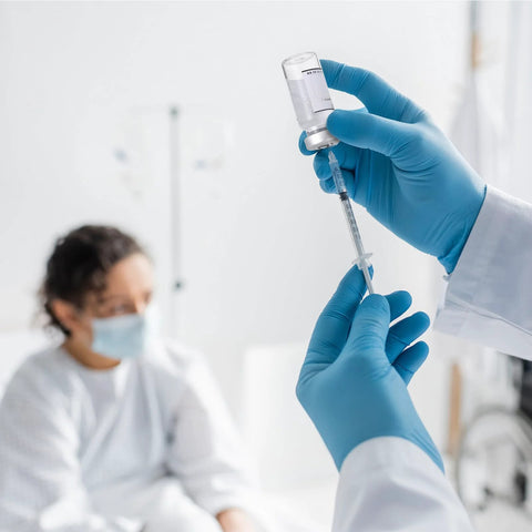 Sealed glass vial being drawn into a syringe by blue-gloved hands in a clinical setting.