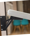Front view of black folding lifting hinge with coil spring mounted to a wooden door frame, supporting a white padded panel.