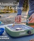 Person handling Cascade 18 1-Burner Camping Stove With 8000 BTU Output And Matchless Lighting, outdoor setup, green aluminized steel cooktop