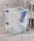 Six Pack Of 66 Quart Clear Plastic Storage Containers With Sleek Blue Latching Lids Designed For Heavy Duty Stackable Organization And Hassle Free Clutter Control In Your Living Room Or Home Office, stacked with pillows and blankets.