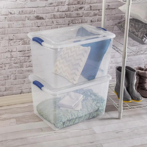 Six Pack Of 66 Quart Clear Plastic Storage Containers With Sleek Blue Latching Lids Designed For Heavy Duty Stackable Organization And Hassle Free Clutter Control In Your Living Room Or Home Office, stacked with pillows and blankets.