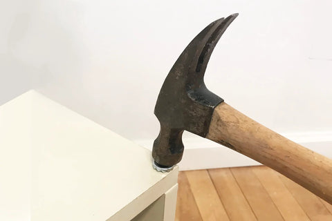 Hammer with a wooden handle removing a nail from a white piece of furniture on a wooden floor.