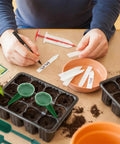 Person using 7 Piece Mini Seed Spreader Tool Set For Precise Seed Dispensing And Easy Garden Planting, labeling plant tags.
