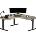 Total Game Changer Electric Height Adjustable Standing Desk With Chill L Shaped Corner Layout, 60 By 60 Inch Executive Rise Workstation For Dual Monitors, Smooth Motor, Black Frame Bright Beech Top in a home office setup.