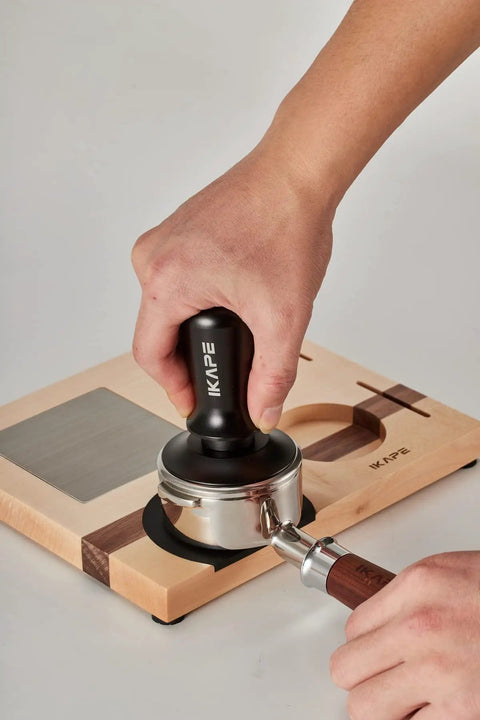 Hand using espresso tamper on portafilter with Espresso Tamper Base And Accessories Organizer In Natural Walnut All Size Portafilter Compatible Tamping Station Holder 4-In-One Coffee Bar Setup Perfect For Home Barista Use And Small Coffee Shops.