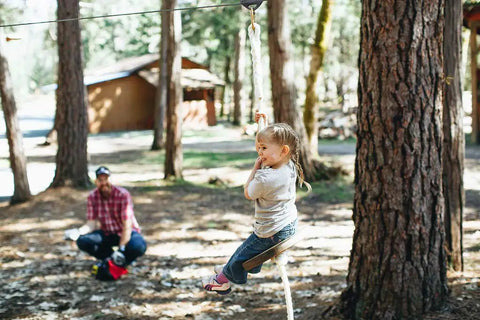 Child riding a Heavy Duty Wooden Zip Line Seat for Backyard Adventures, Pine Wood Frame Holds Up To 350 Lbs, Soft White Rope.