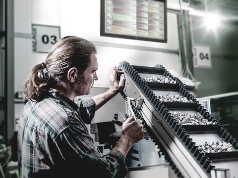 Worker inspecting conveyor belt with metal parts in factory setting, highlighting Zyklop Three Eighths Inch Hex Socket 19mm Size With 29mm Length Ball Intercept Ring Rear End Knurling Chrome Plated Finish Beefy Precision For Manual And Machine Use.
