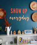 Home office with wall baskets, 'Show Up Everyday' sign, and white shelves containing Fabri Fix Bonding Solution With Fast Grab And Quick Dry For Fabric Leather Suede Felt And Canvas Washable After Set No Heat No Mess Burn Free 8 Ounce Multimaterial Adhesive.