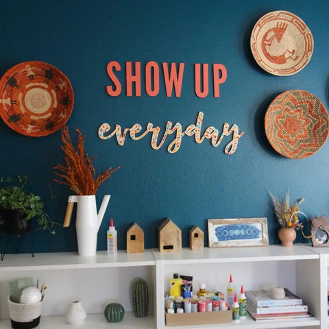 Home office decor with shelves, art supplies, and wall baskets beneath 'SHOW UP everyday' sign on blue wall