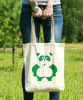 Person outdoors holding a tote bag with a green panda graphic, standing in grass with trees in background, wearing jeans and bracelet.